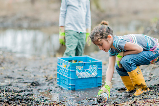 A Caucasian brother and sister are cleaning up garbage in a muddy forest. They are celebrating Earth Day. They are wearing casual clothes, yellow rubber boots, and gloves. The sister is picking up a plastic bottle from the ground.