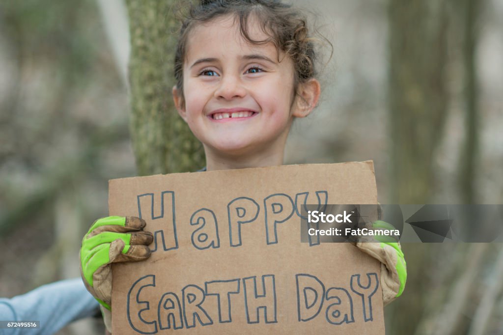 Earth Day A Caucasian girl is celebrating Earth Day. She is wearing casual clothes and gloves. She is smiling and holding up a sign that says "Happy Earth Day". She is in a forest. Earth Day Stock Photo