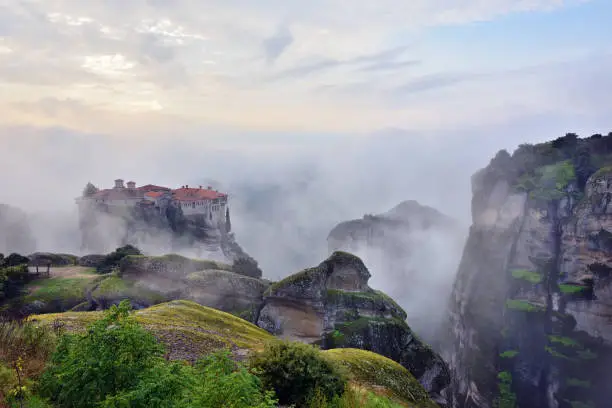 Meteora monasteries. Beautiful view on the Holy Monastery of Varlaam placed on the edge of high rock covered of the morning clouds and mist at sunrise, Kastraki, Greece