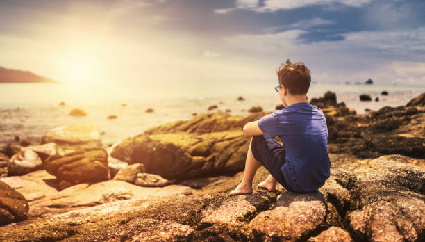 istock Cute boy sitting on rocks and watching at tranquil sea 672559686