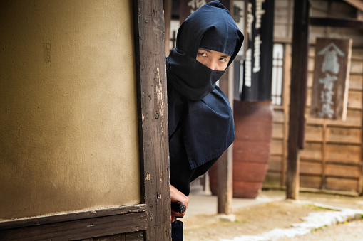 Japanese ninja in black costume hiding in Edo village, peeking around the corner of an old building.