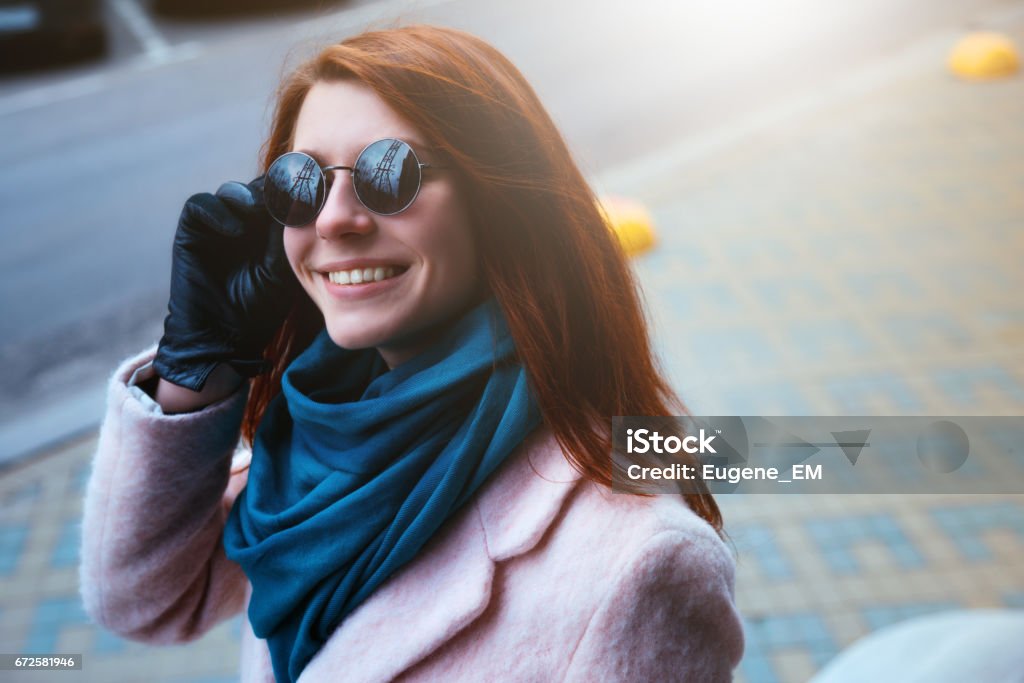 Red haired beautiful girl is walking by the street in a pink coat and blue scarf, with sunglasses. Red haired beautiful girl is walking by the street in a pink coat and blue scarf, with sunglasses. The photo was made on a sunny wonderful day. Adult Stock Photo