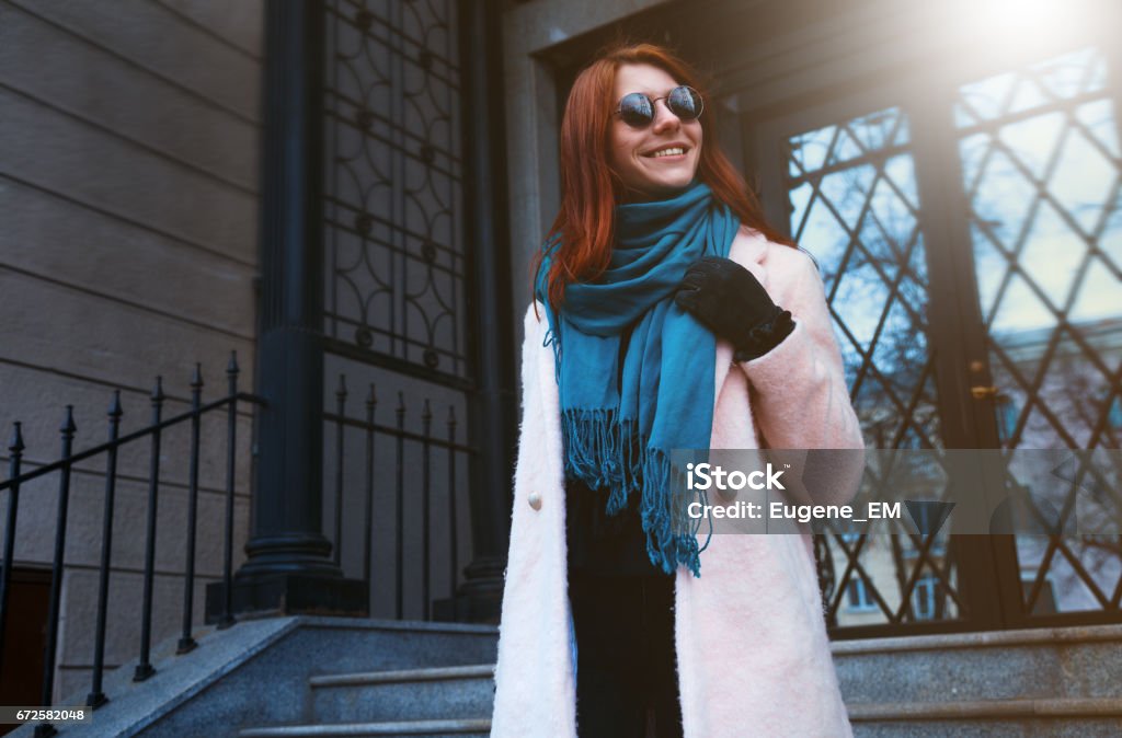 Garota linda de cabelo vermelha está andando pela rua em um casaco cor de rosa e um lenço azul, com óculos de sol. - Foto de stock de Adulto royalty-free