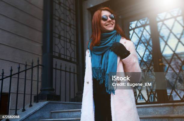 Foto de Garota Linda De Cabelo Vermelha Está Andando Pela Rua Em Um Casaco Cor De Rosa E Um Lenço Azul Com Óculos De Sol e mais fotos de stock de Adulto