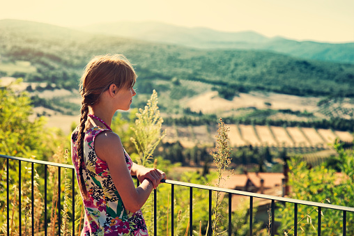 Little girl standing at balcony and looking at tuscan hills view.\n\n