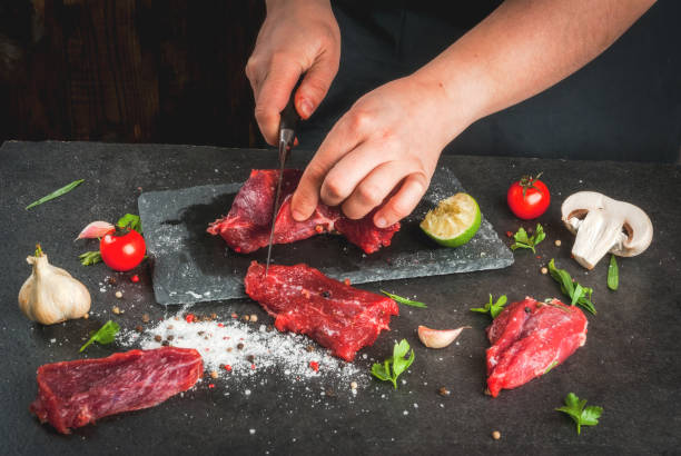 Сooking beef meat Preparation of dinner. Cooking, processing of meat (beef, tenderloin). Person (female hands) cuts a piece of meat into steaks (pieces). Black concrete table, spices and herbs in the frame. tarragon cutting board vegetable herb stock pictures, royalty-free photos & images