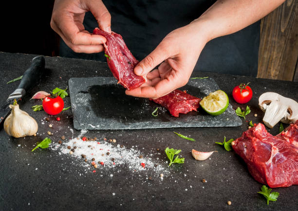 Сooking beef meat Preparation of dinner. Cooking, processing of meat (beef, tenderloin). Person (man's hands) marinates meat steaks. Black concrete table, spices and herbs in the frame. tarragon cutting board vegetable herb stock pictures, royalty-free photos & images