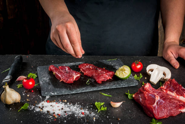 Сooking beef meat Preparation of dinner. Cooking, processing of meat (beef, tenderloin). Person (man's hands) marinates meat - sprinkles with salt, pepper. Black concrete table, spices and herbs in the frame. tarragon cutting board vegetable herb stock pictures, royalty-free photos & images