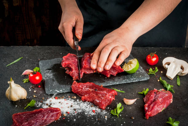 Сooking beef meat Preparation of dinner. Cooking, processing of meat (beef, tenderloin). Person (female hands) cuts a piece of meat into steaks (pieces). Black concrete table, spices and herbs in the frame. tarragon cutting board vegetable herb stock pictures, royalty-free photos & images