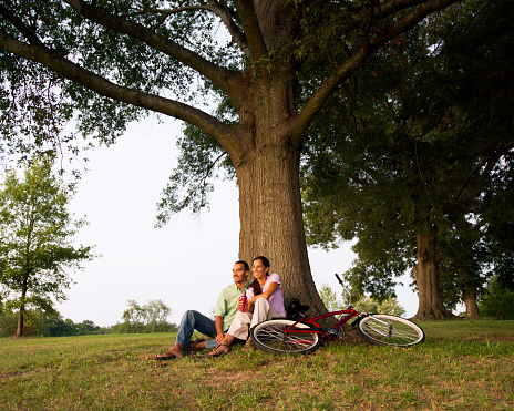 Couple sitting next to large tree with bicycle in the foreground.