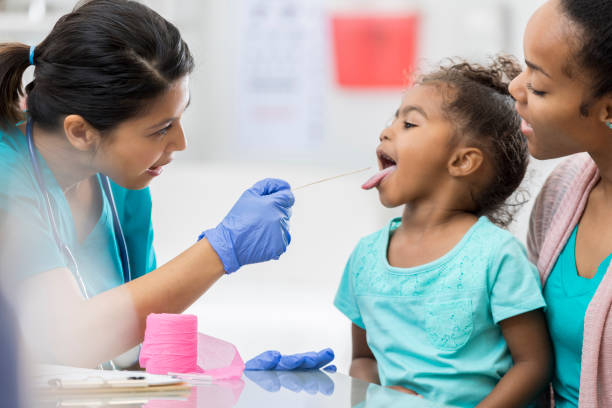 Doctor examines young African American patient Confident mid adult Asian female pediatrician uses a tongue depressor to look at young African American girl's throat. The girl's mother is holding her. emergency medicine stock pictures, royalty-free photos & images