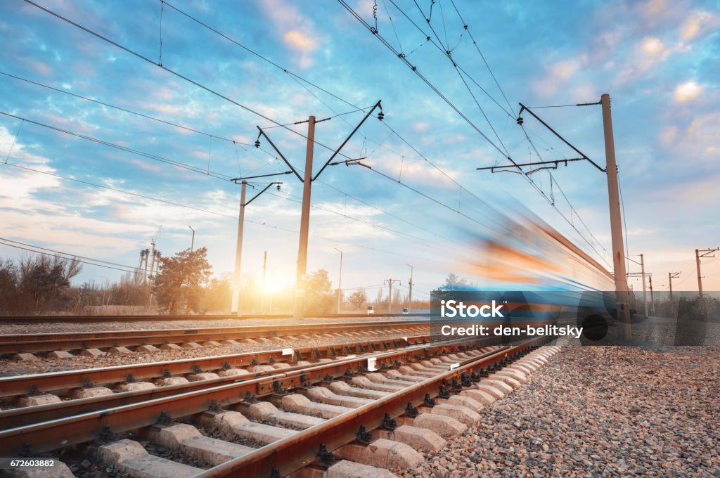 High speed blue passenger train in motion on railroad at sunset. Blurred commuter train. Railway station against colorful sky. Railroad travel, railway tourism. Rural industrial landscape. Vintage Rail Transportation Stock Photo