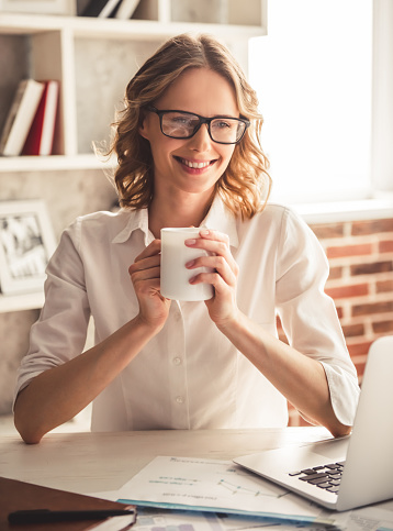 Beautiful business woman in eyeglasses is drinking coffee and smiling while working in office