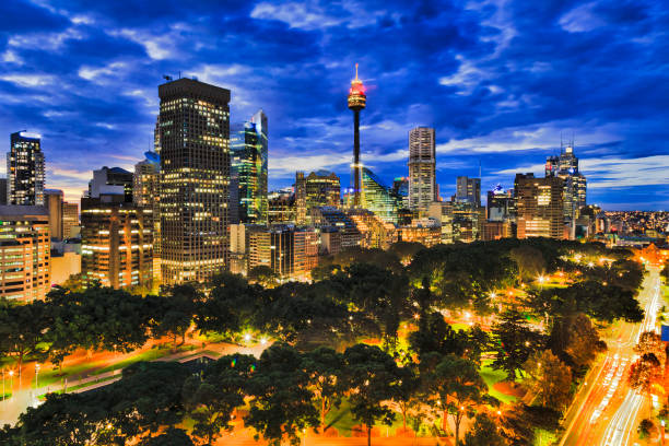 Sy hyde park dark 2 city Inside sunset over Sydney city downtown from Hyde park towards high-rises and towers. Brightly illuminated city architecture between traffic roads under cloudy sky. hyde park sydney stock pictures, royalty-free photos & images