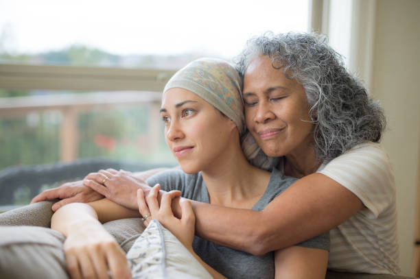 hawaiian woman in 50s embracing her mid-20s daughter on couch who is fighting cancer - cancer chemotherapy drug patient women imagens e fotografias de stock