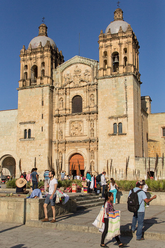 Photograph of several people  in front of the Temple of Santo Domingo de Guzman, in  Oaxaca, Mexico during a sunny day. In front of the baroque façade, some agaves.