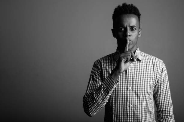 foto de estúdio do jovem empresário africano com cabelo encaracolado, contra um fundo cinza em preto e branco - finger on lips black and white secrecy men - fotografias e filmes do acervo