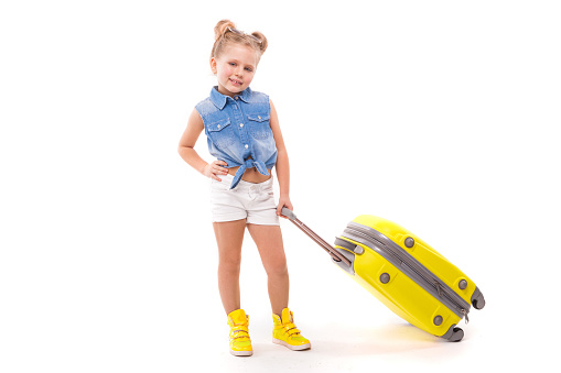 Isolated on white, cute little caucasian blonde girl in blue shirt, white shorts, sunglasses and yellow boots hold yellow suitcase by the handle, hand on waist, look at camera