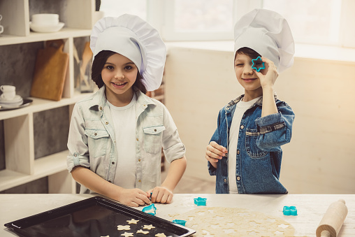 Cute children in chef hats are using cookie cutters and smiling while making cookies in kitchen