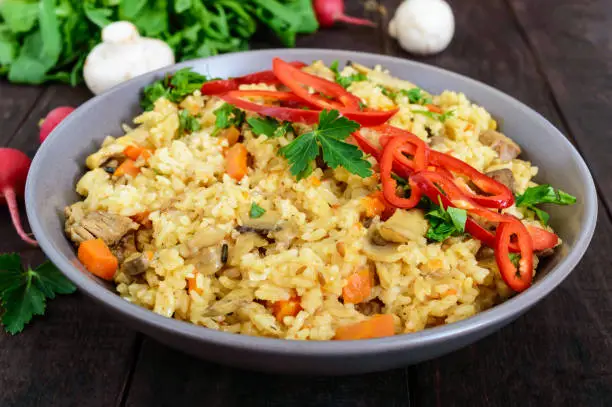 A traditional Asian dish - pilaf with meat, mushrooms and pepper capi in a bowl on a dark wooden background.