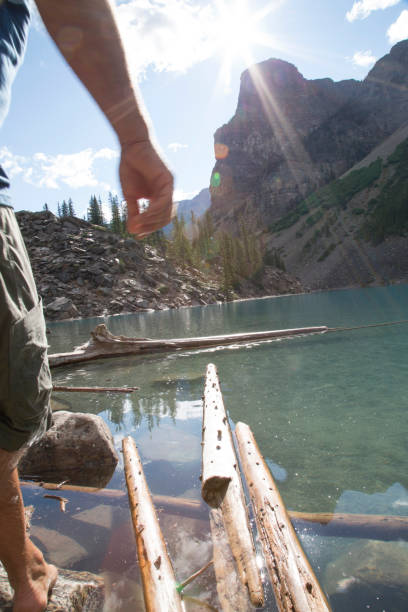 istock Man walks along logs into lake, mountains behind 672817863