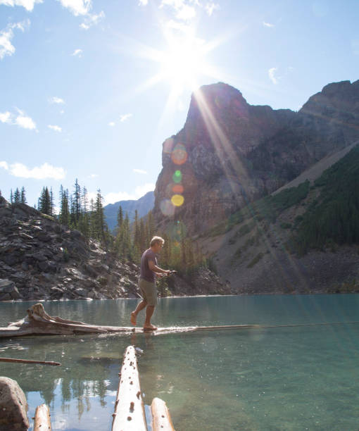 istock Man balances along log into lake, mountains behind 672817873