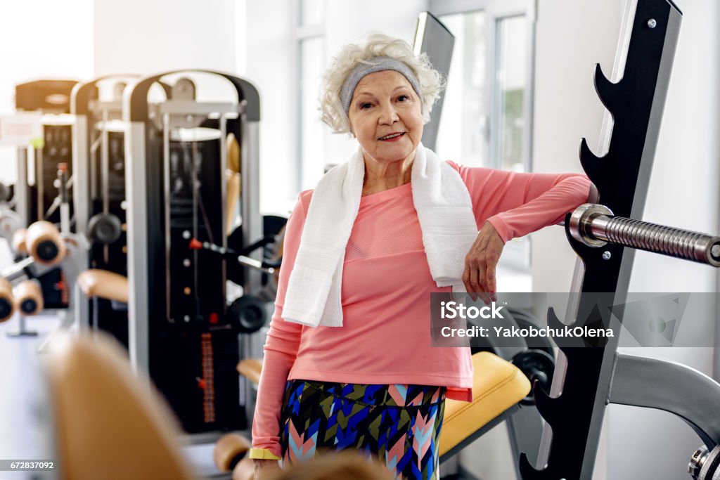 happy grandmother in fitness center Outgoing old woman leaning on tool. She pumping iron in exercise room. Portrait Grandmother Stock Photo