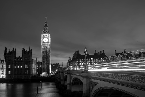 Black and white night shot of light trails on the Westminster Bridge in London and a view of a Big Ben and the Houses of a Parliament.