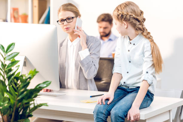 istock daughter sitting on table, businesspeople working with computer behind in office 672923414