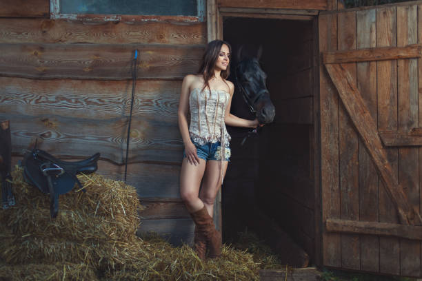 Woman in a Western-style outfit posing with a brown horse in a hay barn. She wears a red plaid shirt, blue jeans, and a brown leather jacket while holding a red cowboy hat. Large hay bales in the background.
