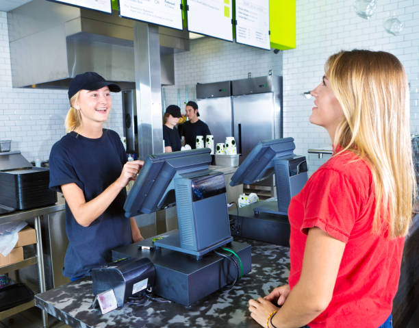 Checkout Server Serving Young Woman Customer Ordering at Fast Food Restaurant A young woman customer placing her order at a fast food convenience restaurant. A young woman server staff is assisting her at the checkout cashier counter with the kitchen staff working in the background. fast food restaurant stock pictures, royalty-free photos & images