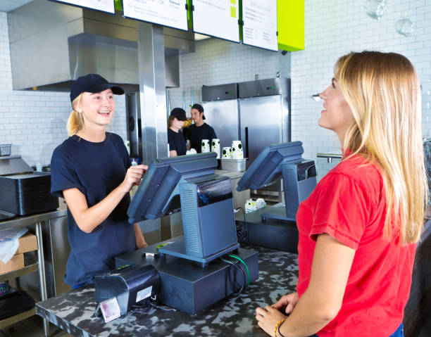 Checkout Server Serving Young Woman Customer Ordering at Fast Food Restaurant A young woman customer placing her order at a fast food convenience restaurant. A young woman server staff is assisting her at the checkout cashier counter with the kitchen staff working in the background. cashier stock pictures, royalty-free photos & images