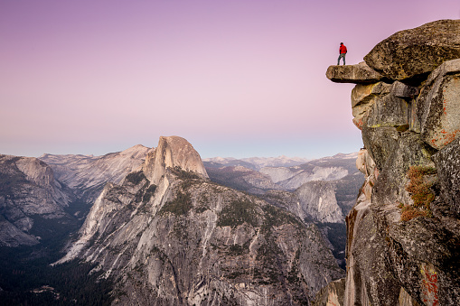 A fearless male hiker is standing on an overhanging rock at Glacier Point enjoying the breathtaking view towards famous Half Dome in beautiful post sunset twilight in summer, Yosemite National Park, California