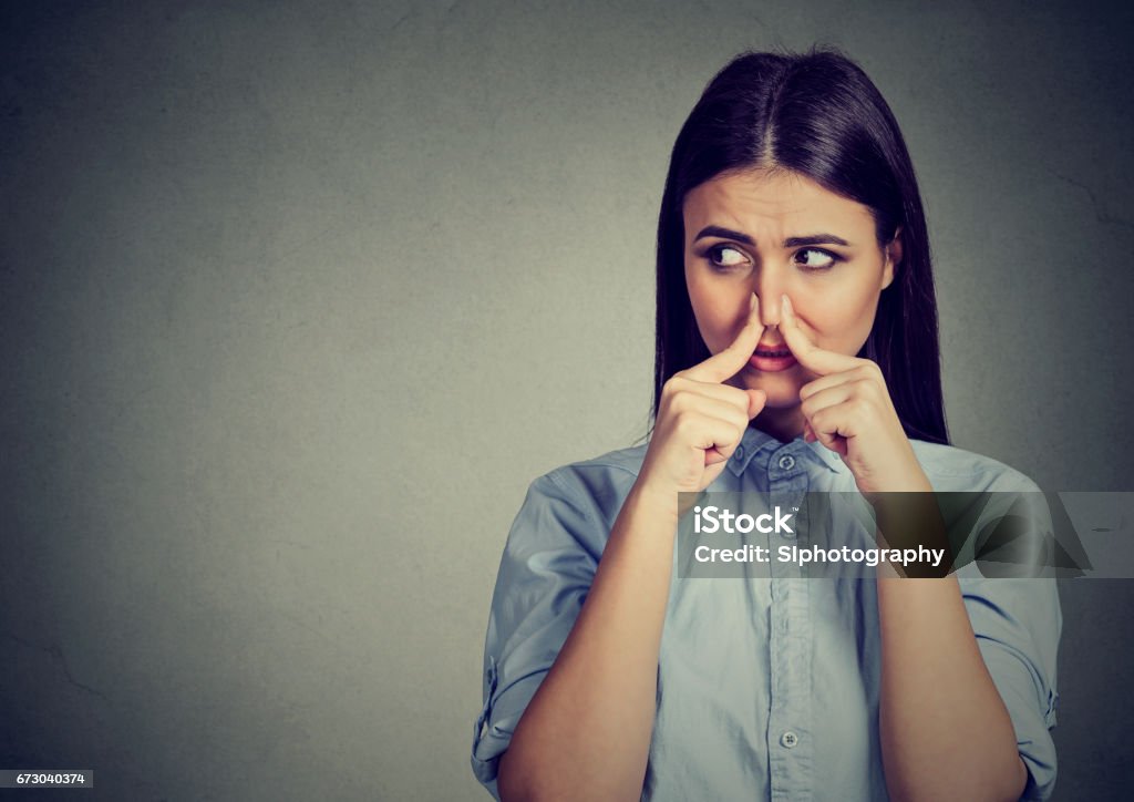 Woman pinches nose with fingers looks with disgust away something stinks bad smell Woman pinches nose with fingers looks with disgust away something stinks bad smell isolated on gray background. Unpleasant Smell Stock Photo