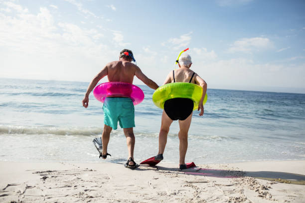 Senior couple in inflatable ring and flipper walking towards sea Rear view of senior couple in inflatable ring and flipper walking towards sea on a sunny day male swimsuit standing arm around stock pictures, royalty-free photos & images