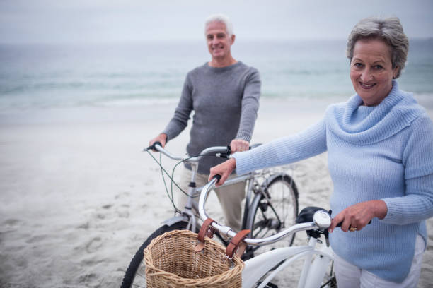 Happy senior couple with their bike Portrait of happy senior couple with their bike on the beach male swimsuit standing arm around stock pictures, royalty-free photos & images