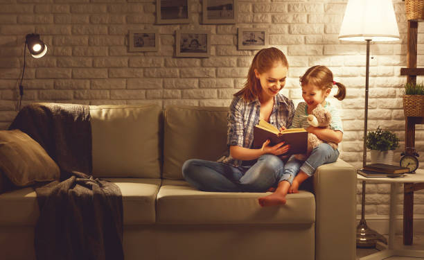 la famiglia prima di andare a letto la madre legge al libro della figlia del bambino vicino a una lampada la sera - bedtime stories foto e immagini stock