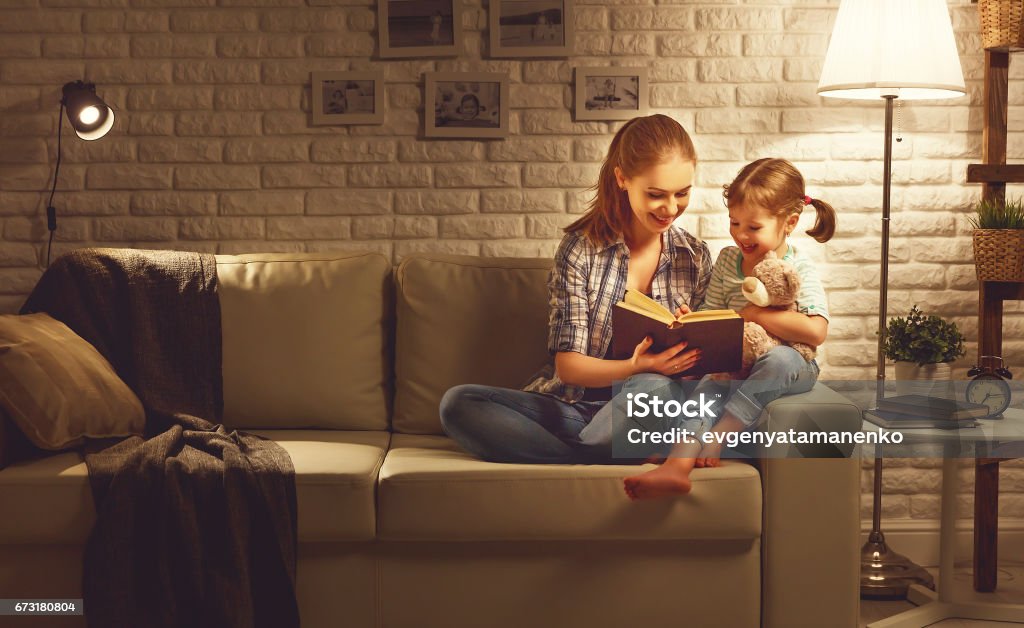 Família antes de ir para a mãe de cama lê ao seu livro de filha de criança perto de uma lâmpada à noite - Foto de stock de Família royalty-free