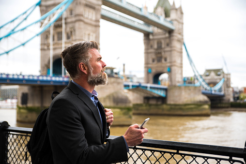 Handsome bearded businessman with a backpack on his shoulder using a smart phone. Tower Bridge in the background. Shot made in London - United Kingdom.