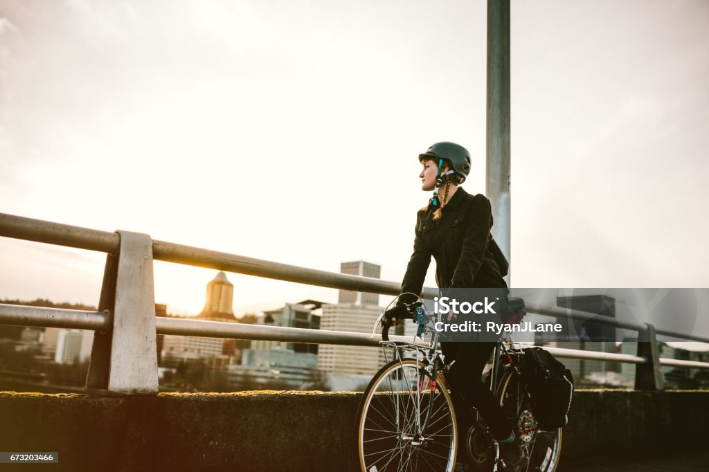 Bike Commuter in Portland Oregon A smiling young woman commuting in an urban city environment on her street bicycle, waterproof panniers on her bike rack. She rides up a ramp from downtown Portland, Oregon, that leads up to the Burnside bridge. Colorful sky from the setting sun. Horizontal image with copy space.  A depiction of young adult / hipster lifestyle in the city. City Stock Photo