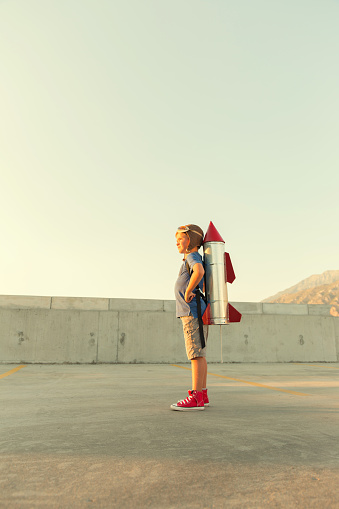 A young boy stands in an urban environment with a homemade rocket strapped to his back. He loves science and technology and wants to fly with a rocket when he grows up.