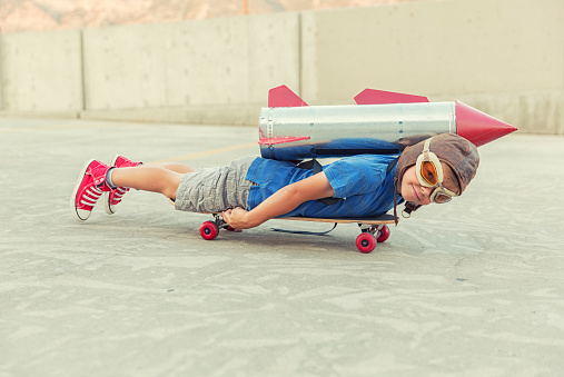 A young boy is lying on a skateboard, in an urban environment with a homemade rocket strapped to his back. He loves science and technology and wants to zoom along in a rocket when he grows up.