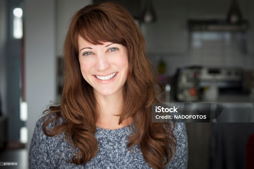 Portrait Of A Mature woman smiling At The Camera. In the kitchen. women, 40s, red hair, fashion model, beauty product, lifestyles, healthy lifestyle, sensuality, copy space, elegance, mature adult, beautiful, cheerful, happiness, smiling, 40-44 years, one person, confidence, female, professional occupation, kitchen, one woman only, leadership, looking at camera, expertise, caucasian ethnicity, occupation, close-up, satisfaction, success, positive emotion, authority, aspirations, adult, toothy smile, global business, cute, achievement, consultant, attractive female, one young woman only, office worker, attitude, staring Women Stock Photo