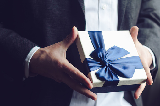 Horizontal close up of Caucasian man in black suit and white shirt offering a gift box with blue large ribbon selective focus natural light