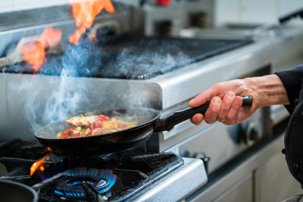 Closeup of the hands of a male chef holding a deep pan and with the other hand a spoon from which a sauce drips.