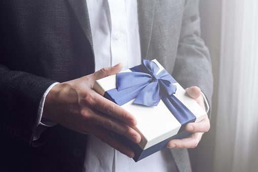 Horizontal close up of Caucasian man in black suit and white shirt offering a gift box with blue large ribbon selective focus low key dark natural light