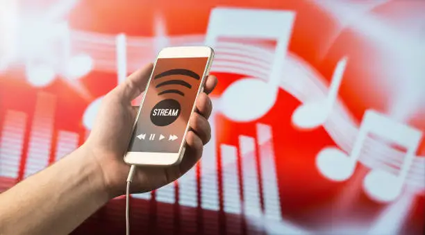 Photo of Modern online music streaming concept. Close up of a man holding smartphone in hand and listening to music with mobile app. Red blurred note background.