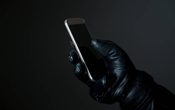 Mobile and cyber security thread concept. Criminal holding smartphone with black leather gloves in darkness. Hacker touching smart phone screen.