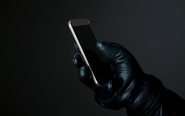 Mobile and cyber security thread concept. Criminal holding smartphone with black leather gloves in darkness. Hacker touching smart phone screen. Modern criminal stealing with cellphone. hoax stock pictures, royalty-free photos & images