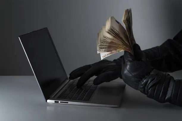 Photo of Internet fraud and cyber security concept. Criminal holding a lot of money and using laptop with black leather gloves.